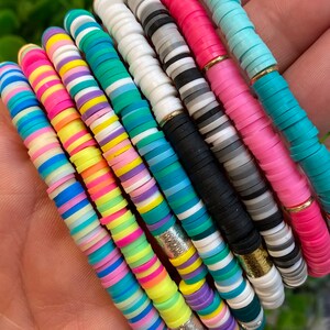 May include: A set of colourful bracelets made of stacked, round, flat beads. The bracelets are in various colours, including pink, blue, green, yellow, and white. Each bracelet has a gold or silver accent.
