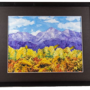 May include: A framed quilted artwork depicting a mountain range with purple peaks and a foreground of yellow and orange trees. The sky is blue with white clouds.