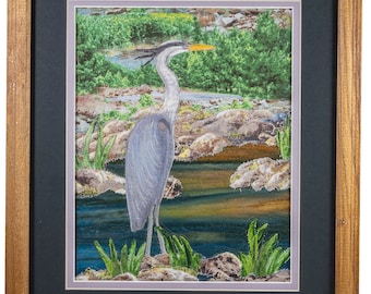 May include: A framed artwork featuring a gray heron standing in a stream with green foliage and rocks. The heron has a long, thin beak and long legs. The background is a blurred image of trees and a stream.