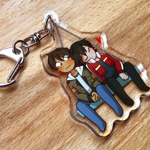 Puede incluir: Un llavero de acrílico transparente con dos personajes de dibujos animados, uno con cabello castaño y una chaqueta verde, el otro con cabello negro y una chaqueta roja, tomados de la mano y sentados en un columpio.