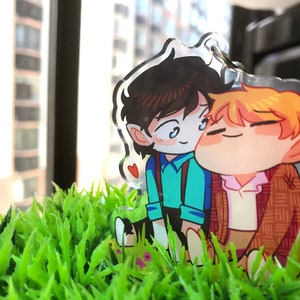 May include: Two cartoon characters, one with brown hair and a blue shirt and the other with blonde hair and a brown jacket, are hugging. They are both smiling and have large eyes. The characters are made of clear acrylic and are sitting on a bed of green grass.