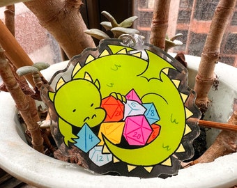 May include: A green cartoon dragon with a sad expression is holding a pile of colorful dice. The dice are numbered 4, 6, 8, 12, 20, and 20. The dragon is on a black background with yellow spikes.