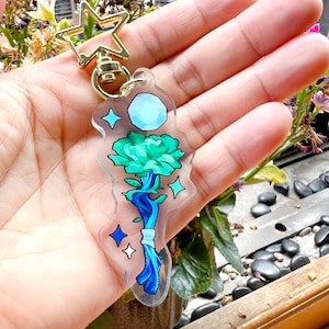 Op de afbeelding: Een heldere acryl sleutelhanger met een goudkleurige stervormige sluiting. De sleutelhanger heeft een bloemenontwerp met een turquoise bloem, blauwe stengel en een lichtblauwe maan. Het ontwerp is geaccentueerd met blauwe sterren en een wit lint.