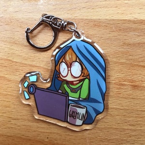 Puede incluir: Un llavero de acrílico transparente con un personaje de dibujos animados que lleva una sudadera con capucha azul, sentado frente a un portátil con una taza que dice "Gremlin".