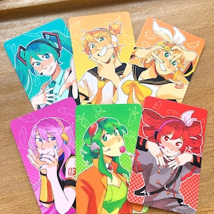 Może przedstawiać: Sześć kolorowych kart z grafikami postaci w stylu anime. Każda karta przedstawia inną postać z unikalnymi włosami i ubraniami. Karty są prostokątne z zaokrąglonymi rogami i ułożone na jasnobrązowej powierzchni.