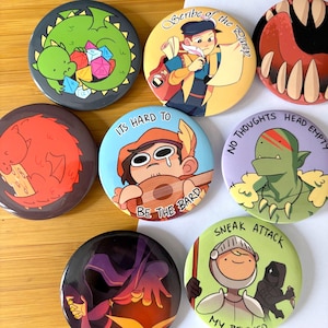 May include: A collection of round, colorful pinback buttons with fantasy-themed illustrations. Designs include a green dragon with dice, a bard with a lute, and a knight with the text "Sneak Attack My Beloved."