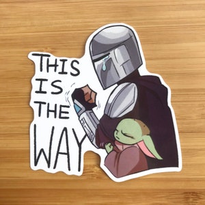 K&ouml;nnte beinhalten: Ein Aufkleber mit einer Cartoon-Illustration eines Mandalorianer-Charakters, der ein kleines, gr&uuml;nes au&szlig;erirdisches Wesen h&auml;lt. Der Text "This is the way" ist in Schwarz auf wei&szlig;em Hintergrund geschrieben.