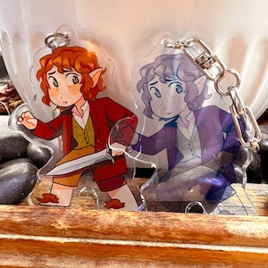 May include: Two cartoon character keychains, one with a red jacket and a sword, the other with a purple jacket. Both characters have pointy ears and are holding swords.
