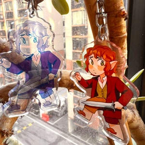 K&ouml;nnte beinhalten: Zwei Schl&uuml;sselanh&auml;nger mit Comicfiguren, einer mit lila Kleidung und einem Schwert, der andere mit einer roten Jacke und einem Schwert. Beide Figuren haben spitze Ohren und halten Schwerter.