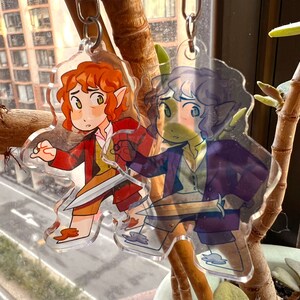 K&ouml;nnte beinhalten: Zwei transparente Acryl-Schl&uuml;sselanh&auml;nger mit Comicfiguren. Der erste Schl&uuml;sselanh&auml;nger zeigt eine Figur mit roten Haaren, einem gr&uuml;nen Hemd und einem Schwert. Der zweite Schl&uuml;sselanh&auml;nger zeigt eine Figur mit lila Haaren, einer lila Jacke und einem Schwert.
