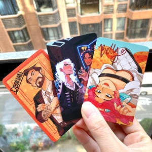 以下が含まれることがあります： スタイリッシュなキャラクターが描かれた3枚のイラストカード。1枚のカードには「Dorian」という名前が書かれています。別のカードには「Betty」という名前が書かれています。3枚目のカードにはネオンサインが背景にあります。カードは窓の前に持たれています。