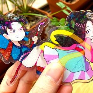 Peut inclure: Deux personnages de style anime sur un ensemble de breloques en acrylique irisé. Le personnage de gauche porte une tenue bleue et noire et tient un bâton rouge et blanc. Le personnage de droite porte une robe rose et bleue avec une ceinture jaune. Les deux personnages ont les cheveux noirs et des accents roses.