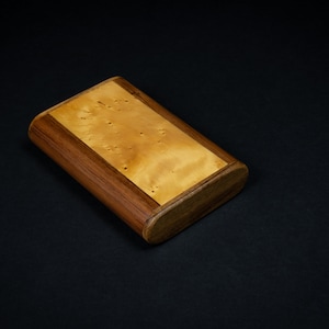 May include: Rectangular wooden box with rounded edges. The box has a light-coloured, speckled wood panel set within a darker wood frame. The box is set against a black background.