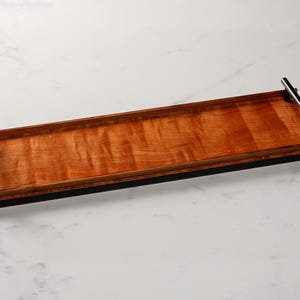 May include: A long, rectangular wooden serving tray with a natural wood grain finish. The tray has two black metal handles on the sides.
