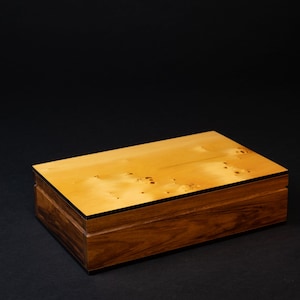 May include: A rectangular wooden box with a light-coloured top and darker wood sides. The top has a speckled pattern and is framed by a thin black border. The box is set against a black background.