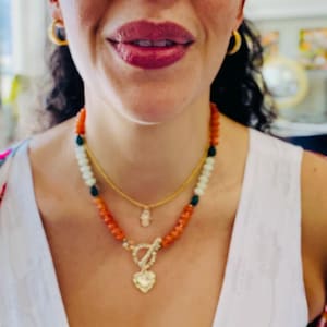 Peut inclure: Un collier à plusieurs rangs avec des perles orange, blanches et vertes, une chaîne dorée et un pendentif en forme de cœur. Le collier est porté avec un haut blanc et des créoles dorées. Un accessoire élégant.
