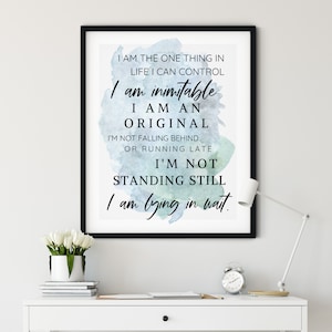 Może przedstawiać: Oprawiony wydruk z czarną obwódką przedstawia inspirujący tekst czcionką w kolorze czarnym na niebiesko-zielonym tle akwarelowym. Wydruk znajduje się na białym biurku z białą lampą, zegarem i wazonem z białymi kwiatami.