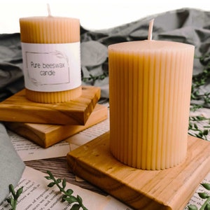 May include: Two light yellow, ribbed beeswax candles. One candle has a label reading "Pure beeswax candle." They are placed on small, square wooden blocks. The background features greenery and a vintage book, creating a natural aesthetic.