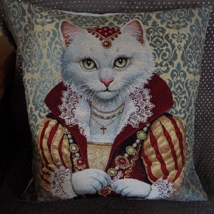 May include: A white cat wearing a red and gold embroidered dress with a white lace collar and a pearl necklace. The cat has a crown and jewels on its head. The background is a blue and gold patterned fabric.