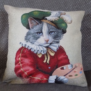 May include: A decorative pillow featuring a gray and white cat wearing a green hat and red jacket, holding a paint palette and brush. The cat has a white ruff around its neck and a yellow bow tie.