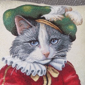May include: A gray and white cat wearing a green and gold hat with a white feather. The cat is wearing a red and gold costume with a white ruffle collar.