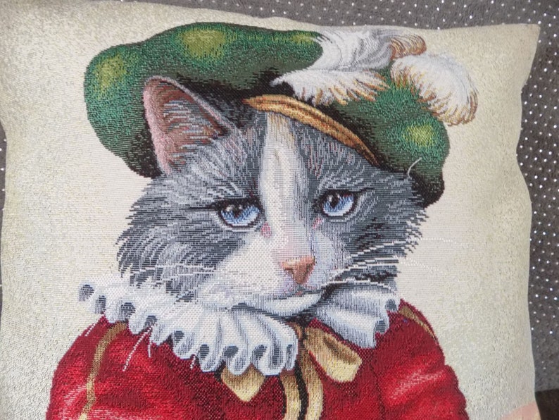 May include: A gray and white cat wearing a green and gold hat with a white feather. The cat is wearing a red and gold costume with a white ruffle collar.