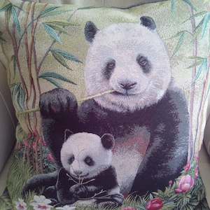 Puede incluir: Una almohada decorativa con un oso panda blanco y negro y una cría de panda más pequeña. La almohada tiene un fondo verde y marrón con plantas de bambú y flores rosas.