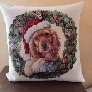 Può includere: Un cuscino decorativo quadrato bianco sporco con un Cocker Spaniel che indossa un cappello da Babbo Natale ed è circondato da una ghirlanda natalizia. La ghirlanda include agrifoglio, bacche, pigne e ornamenti. Il cane tiene in mano un regalo.