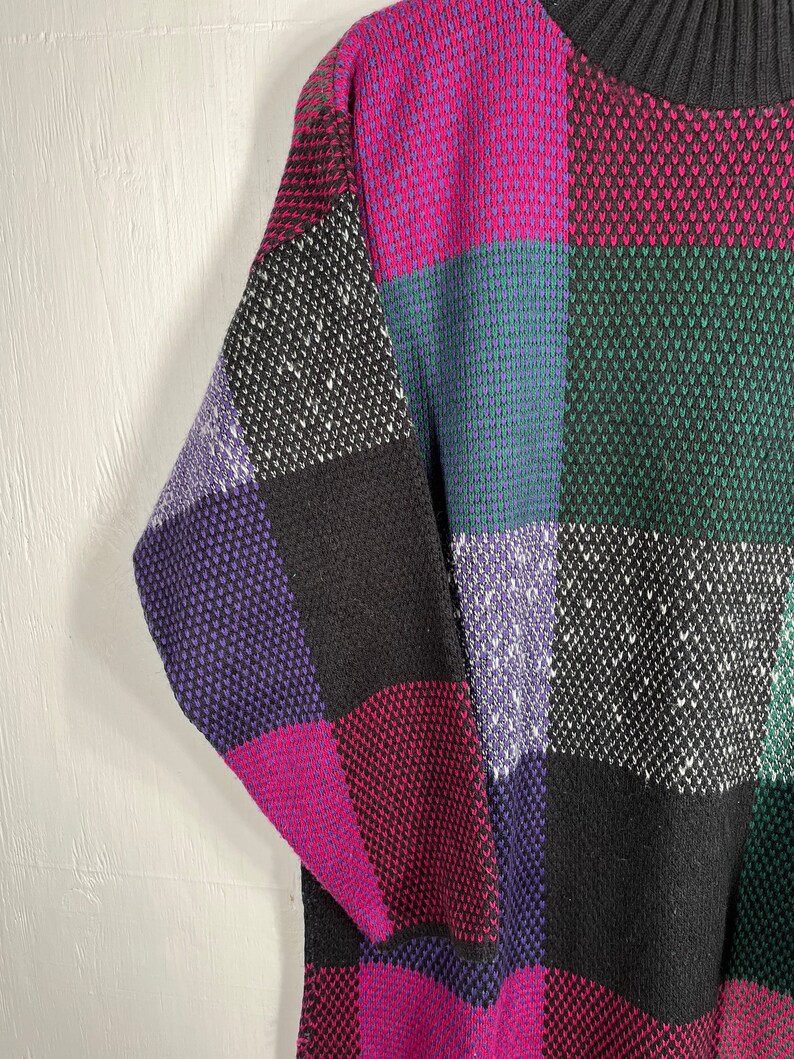 May include: A colorful sweater with a checkerboard pattern. The sweater features squares of black, purple, pink, green, and blue. The sweater has a black ribbed collar and a textured knit design.