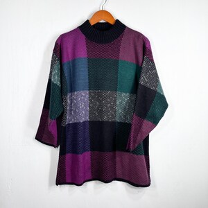 May include: A vintage sweater with a patchwork design in shades of purple, green, blue, and black. The sweater has a mock neck and three-quarter sleeves. It is hanging on a wooden hanger against a white background.