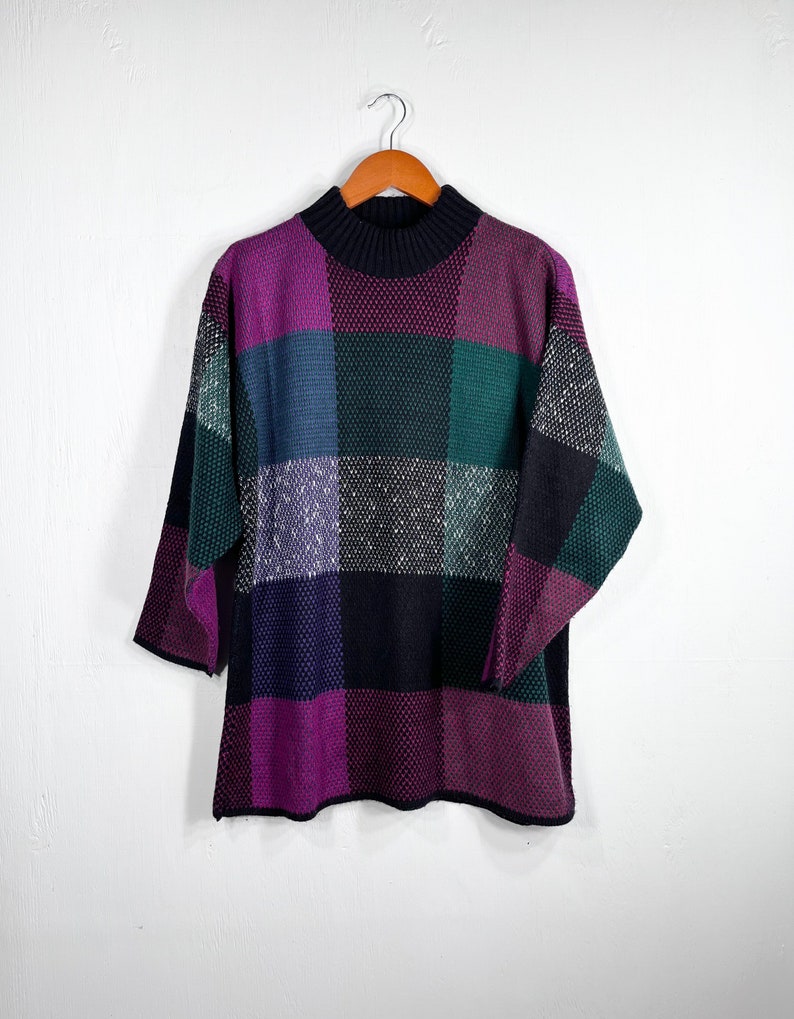 May include: A vintage sweater with a patchwork design in shades of purple, green, blue, and black. The sweater has a mock neck and three-quarter sleeves. It is hanging on a wooden hanger against a white background.