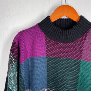 May include: A vintage sweater with a high, ribbed black collar and a colorful patchwork design. The sweater features large squares of magenta, teal, green, and black, creating a bold, geometric pattern. The sweater is hanging on a wooden hanger.