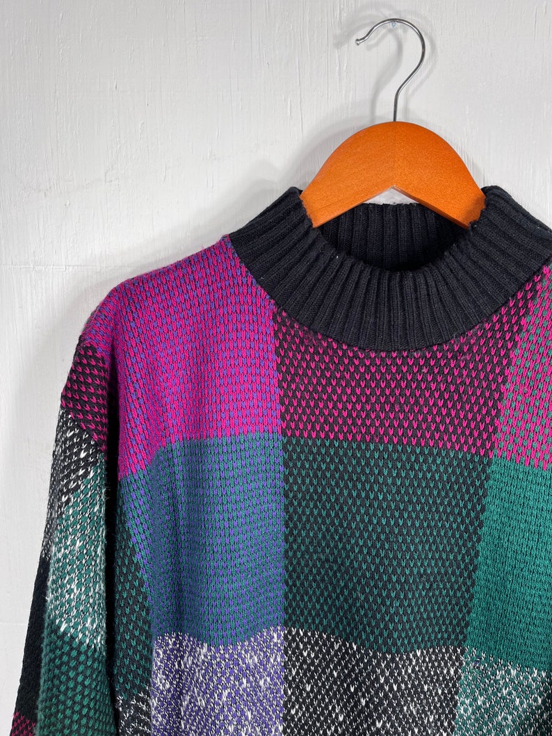 May include: A vintage sweater with a high, ribbed black collar and a colorful patchwork design. The sweater features large squares of magenta, teal, green, and black, creating a bold, geometric pattern. The sweater is hanging on a wooden hanger.