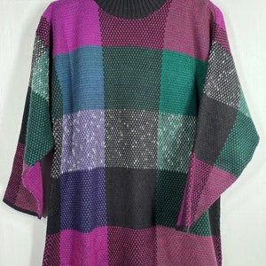 May include: A colorful, oversized sweater with a checkerboard pattern. The sweater features a high, ribbed collar and short sleeves. The squares are in shades of purple, green, black, and white. The sweater is hanging on a wooden hanger.