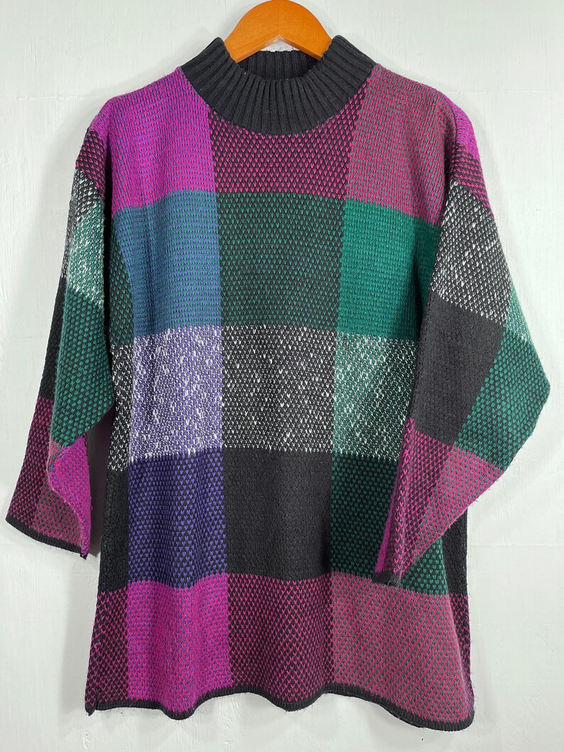May include: A colorful, oversized sweater with a checkerboard pattern. The sweater features a high, ribbed collar and short sleeves. The squares are in shades of purple, green, black, and white. The sweater is hanging on a wooden hanger.