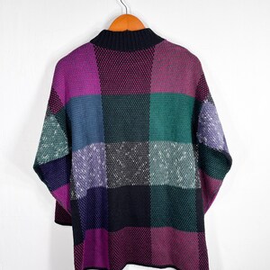 May include: A colorful, patchwork-style sweater with a high, black collar. The sweater features large, square blocks of color, including purple, green, blue, and black. The garment is hanging on a wooden hanger against a white background.