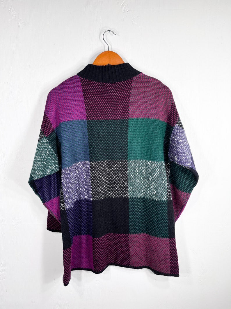 May include: A colorful, patchwork-style sweater with a high, black collar. The sweater features large, square blocks of color, including purple, green, blue, and black. The garment is hanging on a wooden hanger against a white background.
