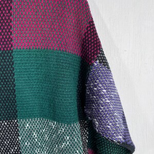 May include: A close-up of a colorful sweater with a patchwork design. The sweater features large blocks of color, including magenta, green, black, white, and purple. The knit fabric has a textured appearance.