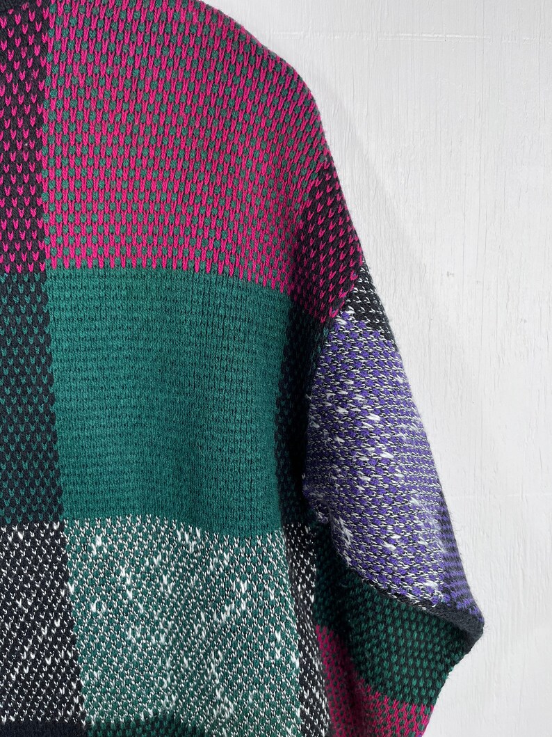 May include: A close-up of a colorful sweater with a patchwork design. The sweater features large blocks of color, including magenta, green, black, white, and purple. The knit fabric has a textured appearance.