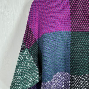 May include: A colorful, patchwork-style sweater in shades of purple, green, and black. The knitted garment features a geometric pattern with a mix of solid and textured blocks. The sleeve is visible, showcasing the design's bold color scheme.