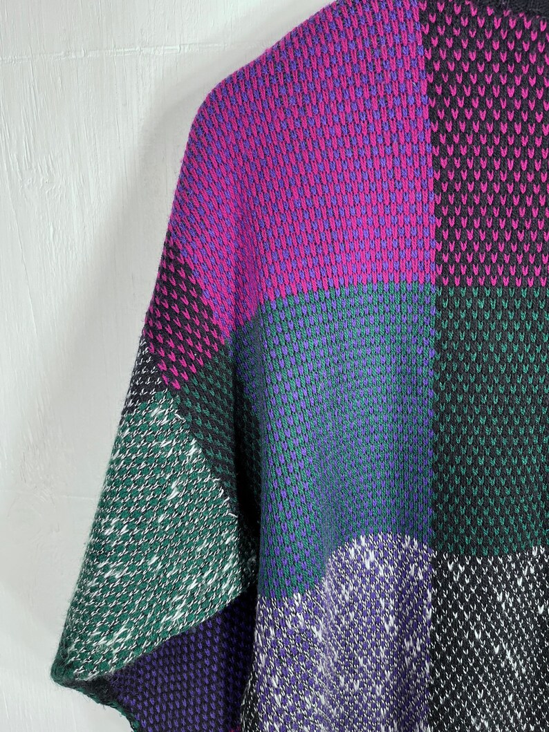 May include: A colorful, patchwork-style sweater in shades of purple, green, and black. The knitted garment features a geometric pattern with a mix of solid and textured blocks. The sleeve is visible, showcasing the design's bold color scheme.