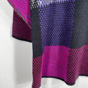 May include: A close-up of a knitted garment with a geometric pattern. The design features blocks of black, purple, magenta, and white. The edges are finished with a green trim. The fabric appears to be thick and textured.
