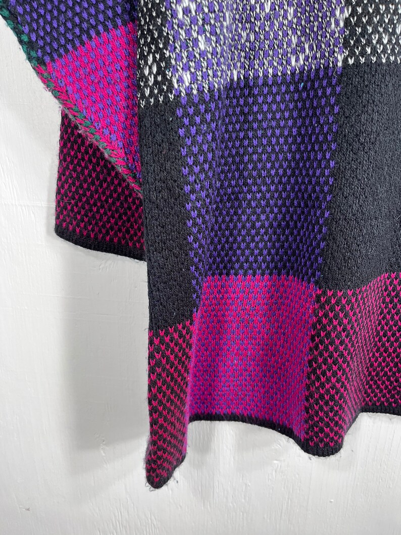 May include: A close-up of a knitted garment with a geometric pattern. The design features blocks of black, purple, magenta, and white. The edges are finished with a green trim. The fabric appears to be thick and textured.