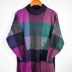 May include: A vintage sweater with a patchwork design in shades of purple, black, green, and blue. The sweater has a mock turtleneck and long sleeves. The sweater is hanging on a wooden hanger against a white background.