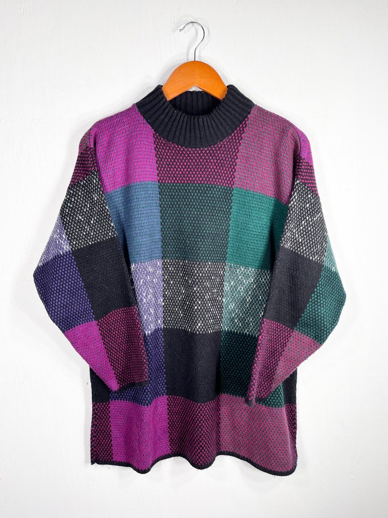 May include: A vintage sweater with a patchwork design in shades of purple, black, green, and blue. The sweater has a mock turtleneck and long sleeves. The sweater is hanging on a wooden hanger against a white background.
