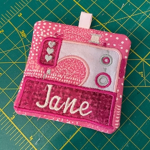 Puede incluir: Cuadrado de tela rosa con un diseño de máquina de coser bordado en rosa y blanco. El nombre "Jane" está bordado debajo de la máquina.