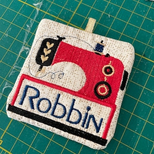 May include: A beige fabric square with a red and black embroidered sewing machine design. The name "Robbin" is embroidered in blue on the bottom of the square.