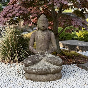 Könnte beinhalten: Eine verwitterte Steinstatue einer sitzenden Figur in meditativer Haltung, die auf einem kreisförmigen Sockel in Lotusform ruht. Die Figur hat einen gelassenen Gesichtsausdruck, die Hände liegen im Schoß und die Oberfläche zeigt Moos.