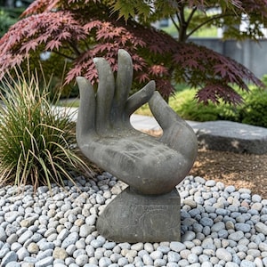 Könnte beinhalten: Eine graue Steinskulptur in Form einer offenen Hand, bei der sich Daumen und Zeigefinger in einer kreisförmigen Geste berühren. Die Skulptur hat eine strukturierte Oberfläche mit geschnitzten Mustern an Basis und Handfläche auf kleinen Steinen.