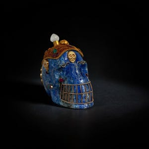 May include: A blue skull sculpture with gold and jewel accents. The skull has a gold crown with small skull details and is adorned with red and green jewels. A white gem sits atop the skull.